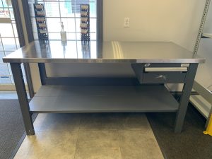 Grey workbench with a stainless steel top, drawer, and lower shelf in an office setting with a key taped to the front of the drawer.